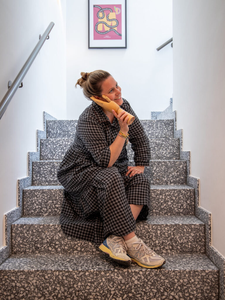 Person sitzt verspielt auf einer Treppe in einem Bürogebäude und hält eine hölzerne Deko-Hand ans Ohr wie ein Telefon; sie trägt ein kariertes Kleid und Sneaker und lächelt zur Seite, im Hintergrund hängt ein farbiges Kunstposter.