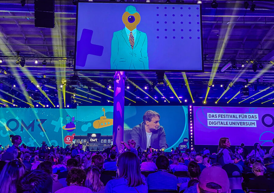 Blick von der Zuschauermenge auf eine grosse Bühne beim OMR-Festival in Hamburg mit bunten Lichtern, Screens und Vortragenden – Team-Ausflug von Compresso.