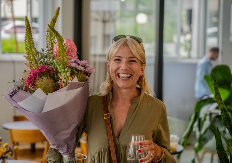 Lächelnde Frau hält einen grossen Blumenstrauss und ein Glas Wein beim Compresso Sundowner 2023.