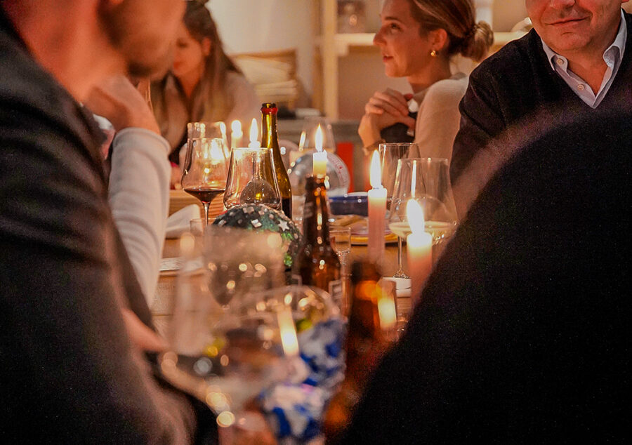 Gemütliches Weihnachtsabendessen 2023: Menschen sitzen an einem festlich gedeckten Tisch mit Kerzenlicht, Wein und Gläsern und führen Gespräche in warmem Ambiente.